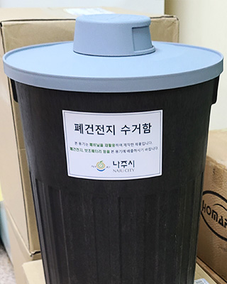 검은색 몸통에 회색 뚜껑으로 세로로 긴 폐건전지 수거함 문구와 나주시 로고가 있는 수거함 모습