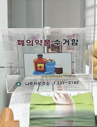 투명하고 직사각형의 플라스틱으로 된 폐의약품 수거함, 나주시보건소 t.339-2142 라고 적혀있는 수거함 정면 모습